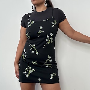 May include: A black mini dress with a white floral pattern, worn over a black short-sleeved top. The dress features thin straps and a fitted silhouette. The floral design includes white flowers and green leaves, creating a contrasting look.