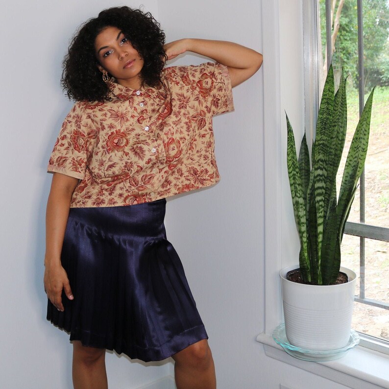 May include: A woman wearing a beige and red floral print cropped shirt with a navy blue pleated skirt.