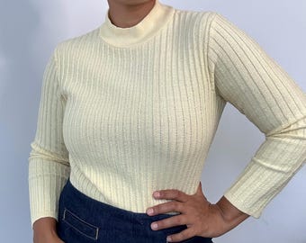 May include: Pale yellow ribbed knit turtleneck sweater with long sleeves. The sweater is paired with dark blue denim pants. The model is wearing red earrings.