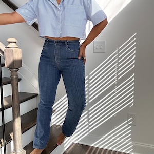 May include: Blue denim jeans with a vertical pinstripe design. The jeans are high-waisted and straight-legged. The model is also wearing a short-sleeved, light blue and white striped button-down shirt.