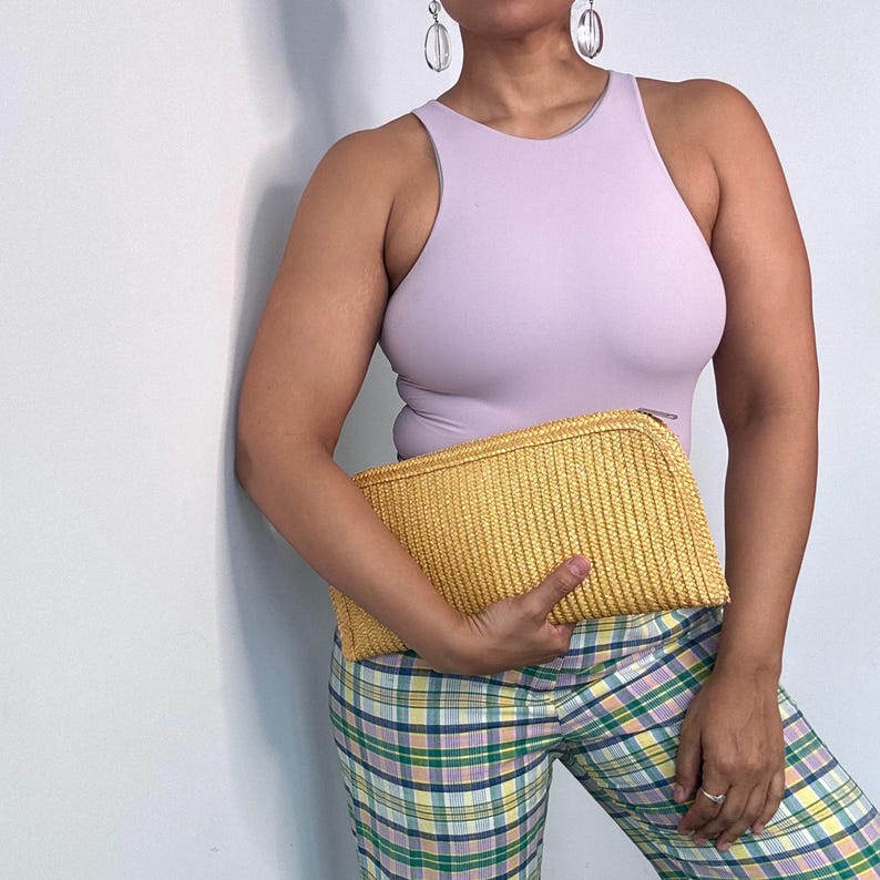 May include: A woman wearing a lavender tank top and plaid pants is holding a yellow woven clutch.