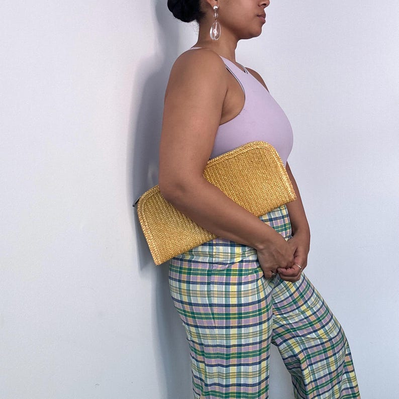 May include: A woman is holding a yellow woven clutch purse. She is wearing a light purple tank top and plaid pants.