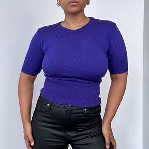 May include: A short-sleeved, royal purple knit top with a ribbed hem and neckline. The top is a solid color and has a fitted silhouette. The model is wearing black pants.