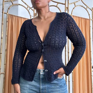 May include: A navy blue, open-weave crochet cardigan with long sleeves and a V-neck. The cardigan has a button-up front with small, round buttons. The person is wearing blue jeans. The background includes a gold-colored metal screen and a peach-colored curtain.