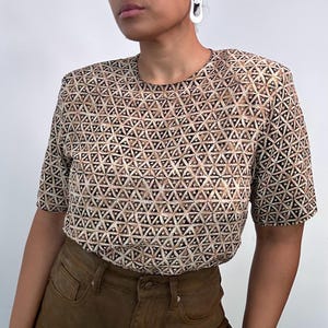 Op de afbeelding: Een top met korte mouwen met een geometrisch patroon in bruin, beige en zwart. Het patroon bestaat uit herhaalde driehoeken. De top wordt gecombineerd met een bruine broek.