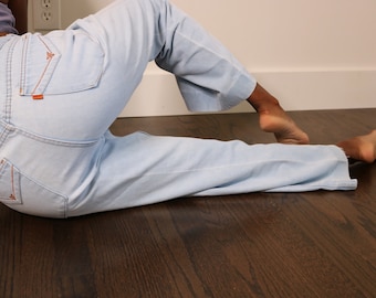May include: A pair of light blue denim jeans with orange stitching. The jeans are lying on a dark wood floor.