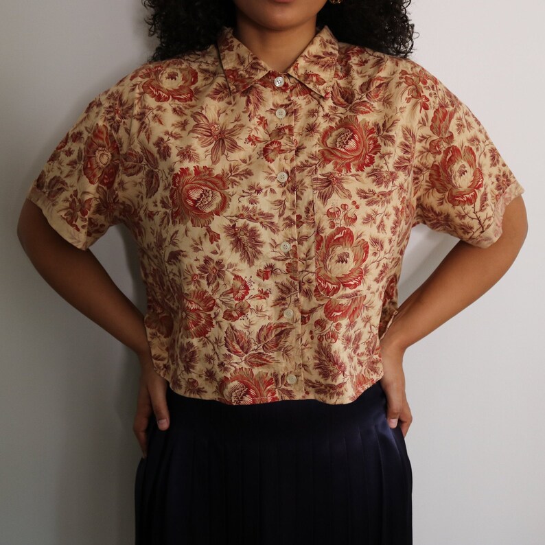May include: A cropped, short-sleeved button-down shirt with a floral print in shades of beige, brown, and red. The shirt has a classic collar and a button-up front.