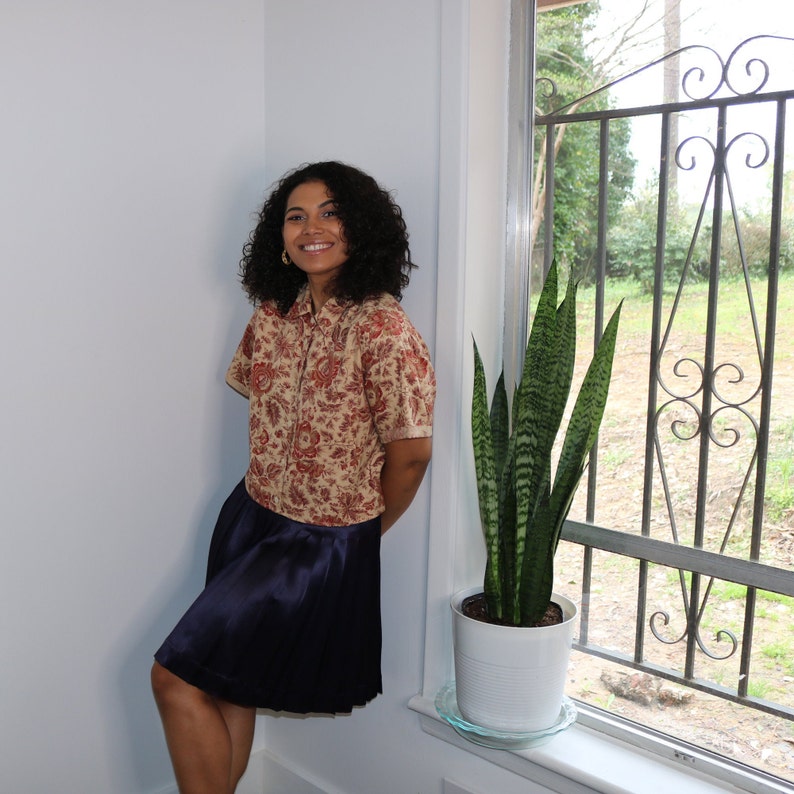 May include: A woman wearing a floral print blouse and a dark blue skirt stands next to a window. A potted snake plant is in front of the window.