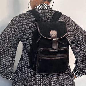 May include: Black suede backpack with brown leather accents. Features a flap closure with a buckle, a drawstring, and a front zippered pocket. The backpack has adjustable straps and a top handle.