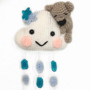 May include: A white crocheted cloud with a smiling face and pink cheeks, with a brown crocheted bear sleeping on top. The cloud has blue and white crocheted raindrops hanging from it.