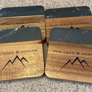 May include: Four square wooden coasters with a black marble top. Each coaster has the text "THE PEAK @ SCHUSS" and a mountain range graphic.