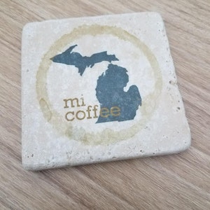 May include: A beige stone coaster with a coffee ring stain. The coaster features a black silhouette of the state of Michigan with the words "mi coffee" in gold lettering.