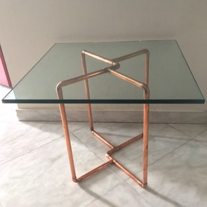 Op de afbeelding: Vierkante bijzettafel met glazen blad en een koperen buizenframe. De tafel heeft een helder glazen blad en een geometrische basis van koperen buizen. De tafel is ongeveer 61 cm vierkant en 51 cm hoog.