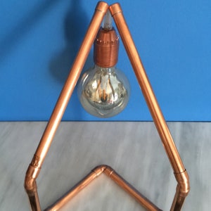 May include: A copper pipe desk lamp with a clear glass bulb. The lamp has a triangular frame and a copper bulb socket. The lamp is on a white surface with a blue background.