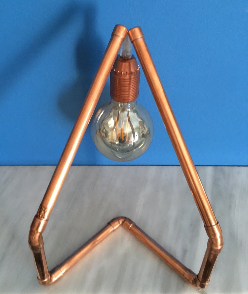 May include: A copper pipe desk lamp with a clear glass bulb. The lamp has a triangular frame and a copper bulb socket. The lamp is on a white surface with a blue background.