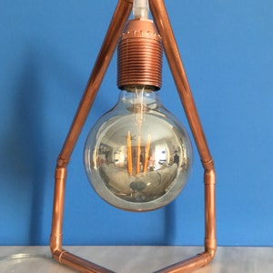 May include: A copper-colored, geometric table lamp with a large, clear, round light bulb. The bulb reflects the surrounding environment. The lamp's frame is made of copper pipes, and the base is a triangle shape.