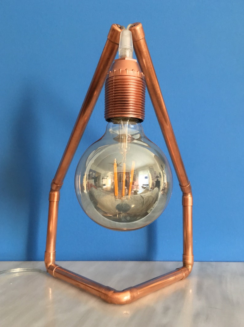 May include: A copper-colored, geometric table lamp with a large, clear, round light bulb. The bulb reflects the surrounding environment. The lamp's frame is made of copper pipes, and the base is a triangle shape.
