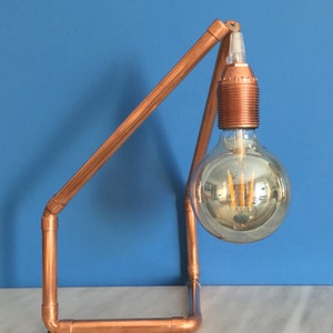 May include: A copper pipe desk lamp with a large, round, clear lightbulb. The lamp has a minimalist design with a rectangular base and an angled arm. The lightbulb has a warm, yellow glow. The background is a solid blue.