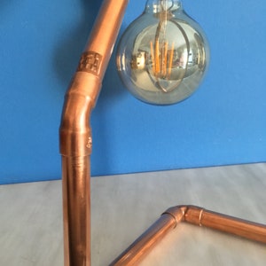 May include: Copper pipe desk lamp with a large, round, clear light bulb. The lamp has a modern, industrial design with a blue background. The bulb has a warm, orange glow.