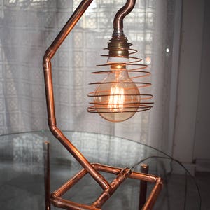 May include: A handcrafted desk lamp made from copper pipes, featuring a vintage-style Edison bulb encased in a spiral wire cage. The lamp's design incorporates copper piping and fittings, offering an industrial and contemporary look.