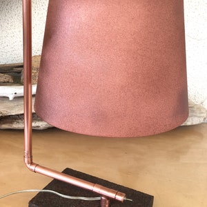 May include: A copper-toned desk lamp with a cylindrical lampshade. The lamp features a copper pipe frame and a rectangular base. The lampshade is a muted rose color. The lamp is designed with a modern, industrial aesthetic.