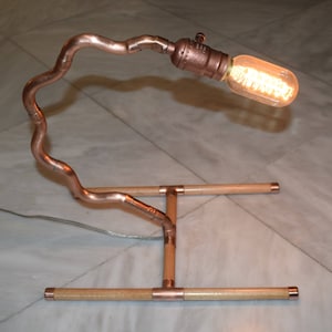Puede incluir: Lámpara de escritorio artesanal de tubería de cobre, con una bombilla Edison vintage. La lámpara presenta un brazo ondulado de cobre y una base de madera con accesorios de cobre. La bombilla está encendida, emitiendo una luz cálida.