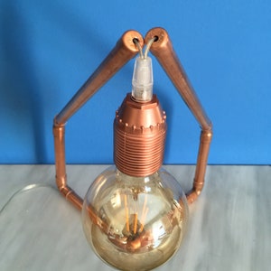 May include: A copper-colored, industrial-style table lamp. The lamp features a large, round, Edison-style light bulb with visible filaments. The bulb is held in place by a copper-colored socket and supported by copper pipes bent into an arch.