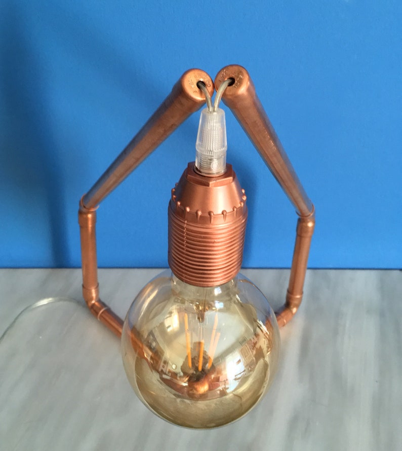May include: A copper-colored, industrial-style table lamp. The lamp features a large, round, Edison-style light bulb with visible filaments. The bulb is held in place by a copper-colored socket and supported by copper pipes bent into an arch.