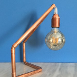 May include: A copper pipe desk lamp with a large, round Edison-style light bulb. The lamp has a geometric design with a base and an angled arm supporting the bulb. The lamp is on a white surface with a blue background.