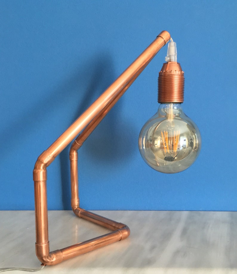 May include: A copper pipe desk lamp with a large, round Edison-style light bulb. The lamp has a geometric design with a base and an angled arm supporting the bulb. The lamp is on a white surface with a blue background.