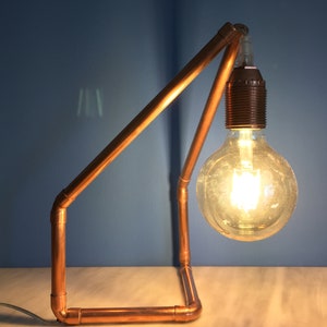 May include: A copper pipe desk lamp with a large, round Edison-style light bulb. The lamp has a geometric frame and a black bulb socket. The light is on, illuminating the bulb and the surrounding area.