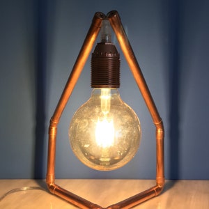 May include: A copper pipe desk lamp with a large, round Edison bulb. The lamp has a geometric frame and a black bulb socket. The bulb is lit, emitting a warm, inviting glow. The lamp is set against a blue background.