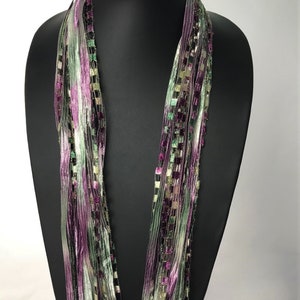 May include: A multi-strand infinity scarf made of thin, silky ribbons in shades of green, purple, and white. The scarf is displayed on a black mannequin.