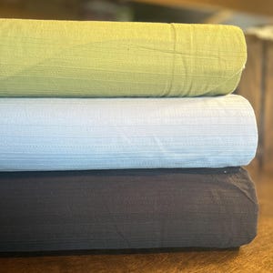 May include: Three rolls of fabric in shades of green, light blue, and dark brown. The fabric has a textured, striped pattern. The rolls are stacked on top of each other, showcasing the different colors and textures of the material.