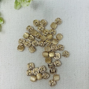 May include: A collection of small, square buttons with a gold-colored metal frame and a light beige, rounded center. The buttons are scattered on a white, textured surface, suggesting a craft or sewing project.