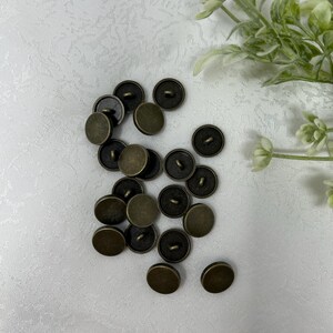 May include: A collection of antique brass-colored buttons scattered on a white textured surface. The buttons are round with a slightly domed shape and a loop on the back for sewing. The buttons have a vintage aesthetic.