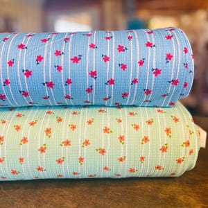 Puede incluir: Dos rollos de tela, uno azul claro y otro verde claro, cada uno con rayas blancas verticales y pequeños diseños florales rojos y rosas. La tela tiene una apariencia texturizada y acolchada, lo que sugiere un material suave y cómodo.