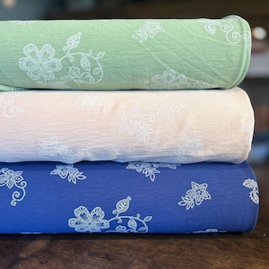 Peut inclure: Trois rouleaux de tissu vert, crème et bleu, chacun orné d'un motif floral blanc. Le tissu est empilé, présentant les différentes couleurs et motifs. Les rouleaux reposent sur une surface en bois.