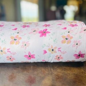 May include: Rolled fabric with a floral design. The pattern showcases pink, yellow, and orange flowers with green leaves against a white backdrop. The material appears to be cotton or a similar fabric.