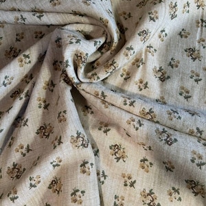 May include: A light beige fabric featuring a repeating pattern of small, muted yellow flowers with dark green stems and leaves. The textile has a visible woven texture and is draped to create soft folds that highlight the floral print design.