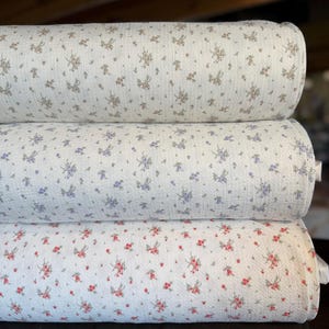 Op de afbeelding: Drie opgestapelde rollen textiel met een textuur en kleine bloemmotieven. De bovenste rol heeft bruine en groene bloemen, de middelste rol heeft paarse en groene bloemen en de onderste rol heeft rode en groene bloemen op een witte achtergrond.