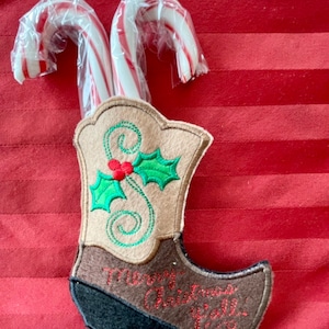 Puede incluir: Un porta calcetines de bota vaquera de fieltro marrón con bordado de acebo verde y el texto "Merry Christmas Y'all". Dos bastones de caramelo rojo y blanco están en la bota.