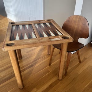 Può includere: Un tavolo da gioco quadrato in legno con tavoliere da backgammon integrato con punte triangolari color crema, marrone e nero, quattro portabicchieri agli angoli e piccole aree rettangolari incassate, accompagnato da una sedia in legno curvo.