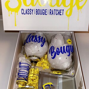 May include: A white gift box with the word "Savage" in yellow and blue dripping font. Inside are two wine glasses with "Classy" and "Bougie" in blue, a shot glass, and a mirrored tray with "Savage" on it.