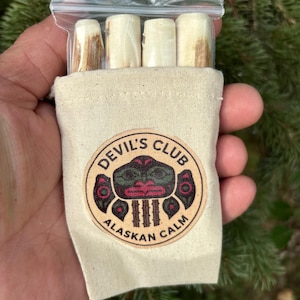 Può includere: Una piccola borsa di tela con la scritta "DEVIL'S CLUB ALASKAN CALM" e una grafica circolare. La borsa contiene diversi oggetti cilindrici di colore chiaro in un sacchetto di plastica trasparente. Lo sfondo è una vista sfocata di fogliame verde.