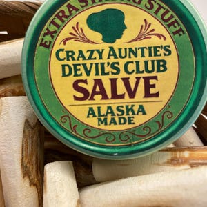 May include: A round green metal tin of Crazy Auntie's Devil's Club Salve, labelled Extra Strong Stuff and Alaska Made, featuring a dark green silhouette of a person's head, resting atop a collection of light-coloured, woody, cylindrical sticks.
