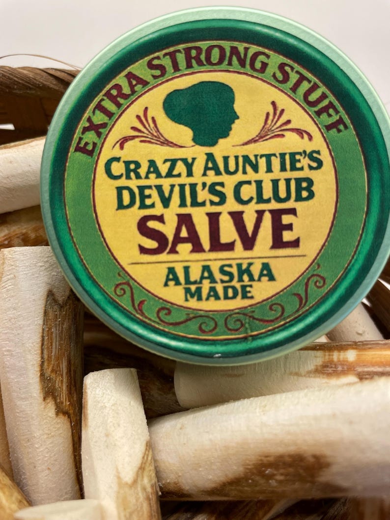 K&ouml;nnte beinhalten: Eine runde gr&uuml;ne Metalldose mit Crazy Auntie's Devil's Club Salve, beschriftet mit Extra Strong Stuff und Alaska Made, mit einer dunkelgr&uuml;nen Silhouette eines Kopfes, die auf einer Ansammlung heller, h&ouml;lzerner, zylindrischer St&auml;bchen liegt.
