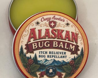 Könnte beinhalten: Eine runde Metalldose mit einem hellgrünen Balsam. Der Deckel trägt den Text Crazy Auntie's ALASKAN BUG BALM, ITCH RELIEVER BUG REPELLANT und 100% NATURAL. Das Design zeigt eine Berglandschaft, Kiefern und eine kleine Karte von Alaska.