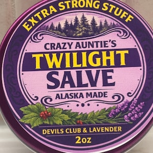 Könnte beinhalten: Eine runde violette Dose mit Crazy Auntie's Twilight Salve, Extra Strong Stuff, Alaska Made, Devils Club & Lavender, 57g. Das Etikett zeigt eine Waldsilhouette, einen dekorativen Rand sowie Illustrationen von grünen Blättern, roten Beeren und Lavendel.