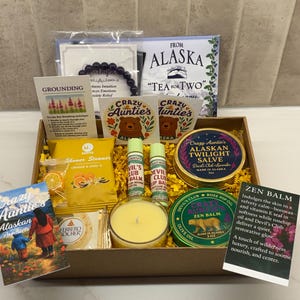 May include: A gift box containing various items including a purple beaded bracelet, a tea packet labeled From Alaska, two lip balms, a yellow candle, a Ferrero Rocher chocolate, a shower steamer, and two round tins labeled Alaskan Twilight Salve and Zen Balm.